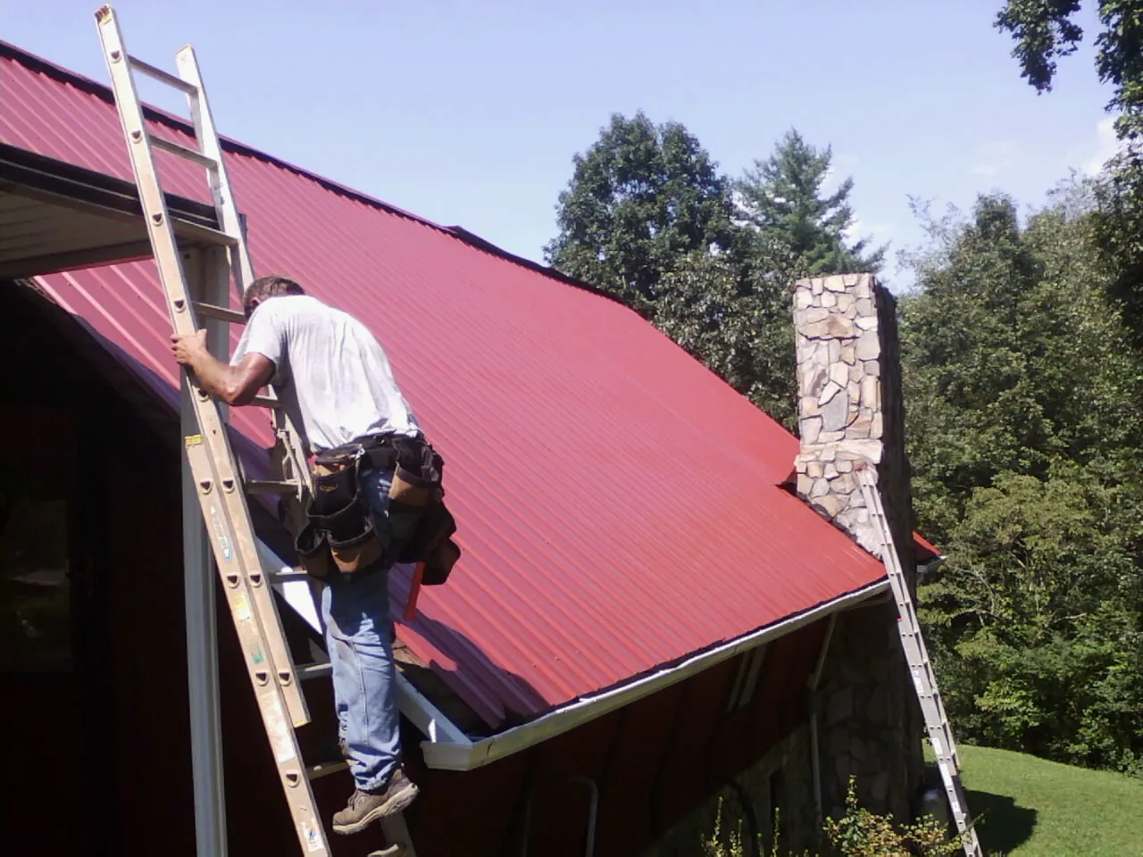 Roofer on a ladder beside a red metal roof and stone chimney — Soffit & Fascia Repair by Roofing Brothers in Libertytown